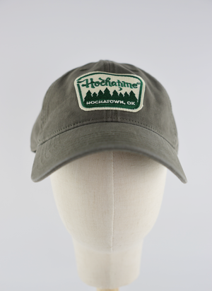 An olive green baseball cap displayed on a cream-colored mannequin head against a light gray background. The front of the cap features an embroidered patch with the text 'Hochaline HOCHATOWN, OK' and a green pine tree silhouette.